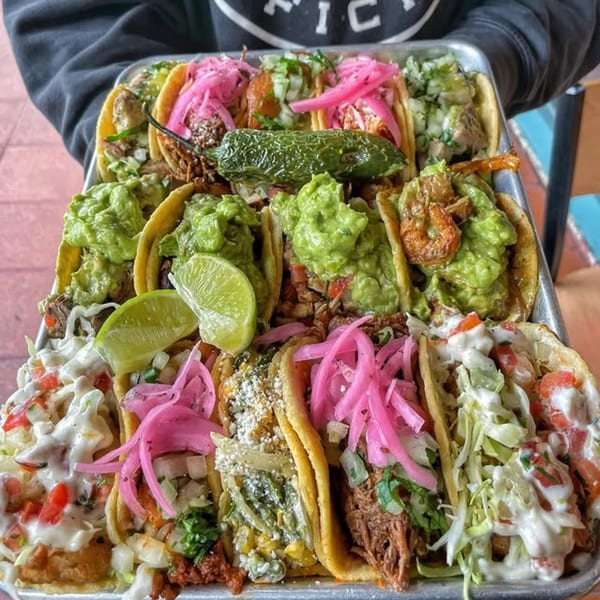 A person holds a large metal tray filled with various La Chancla Mexican Grill tacos, including guacamole and pickled onions.