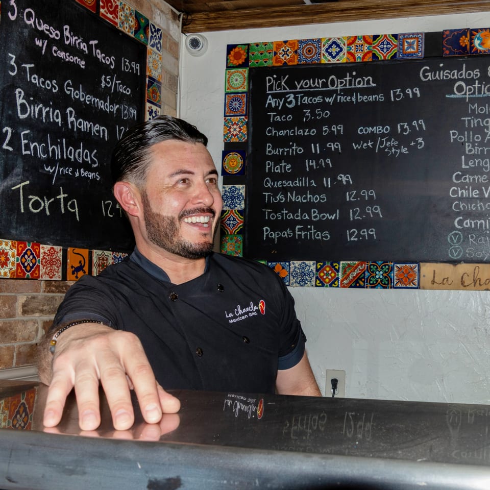 A smiling La Chancla Mexican Grill chef leans on a counter with chalkboards listing menu items.