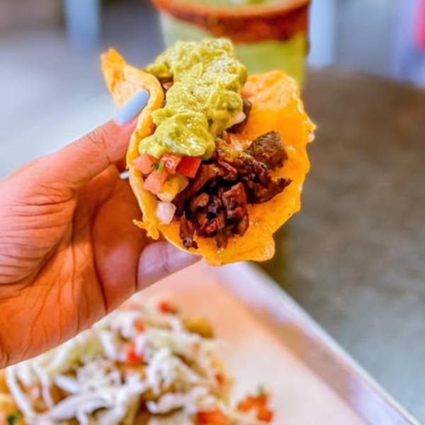 A hand holds a La Chancla Mexican Grill taco with meat, pico de gallo, and guacamole.