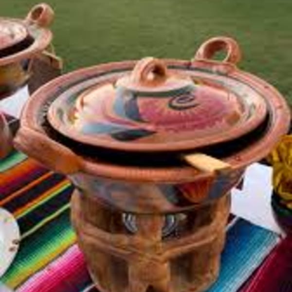 A traditional Mexican clay pot with a colorful lid sits on a wooden stand, on a striped serape.