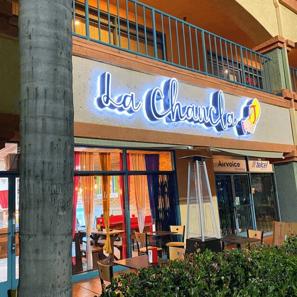 Exterior of La Chancla Mexican Grill at night, with a glowing blue neon sign and outdoor seating.