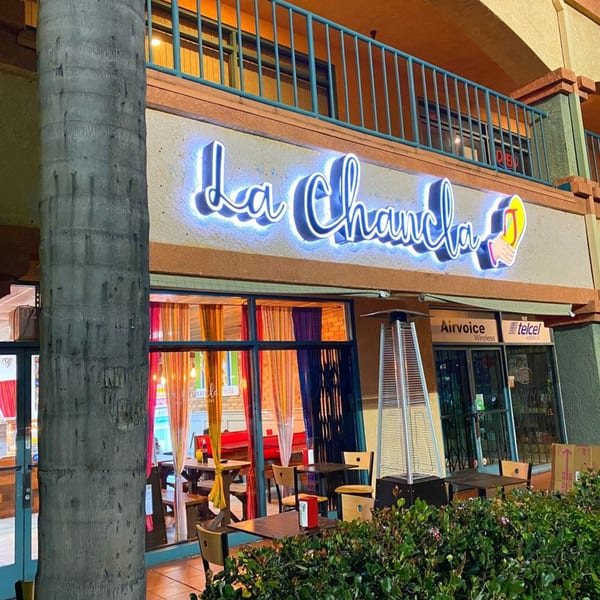 Exterior of La Chancla Mexican Grill at dusk, with a glowing neon sign and outdoor seating.