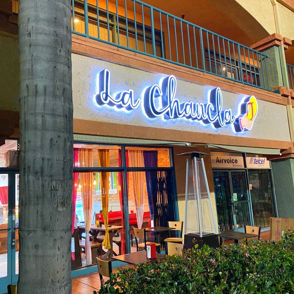 Exterior of La Chancla Mexican Grill at dusk, with a glowing neon sign and outdoor seating.