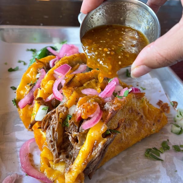 Hand pouring salsa from a metal cup onto three birria tacos with pickled onions and cilantro.