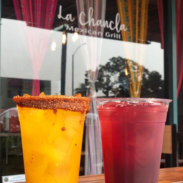 Two drinks, one yellow with a chili rim and one red, in front of La Chancla Mexican Grill.