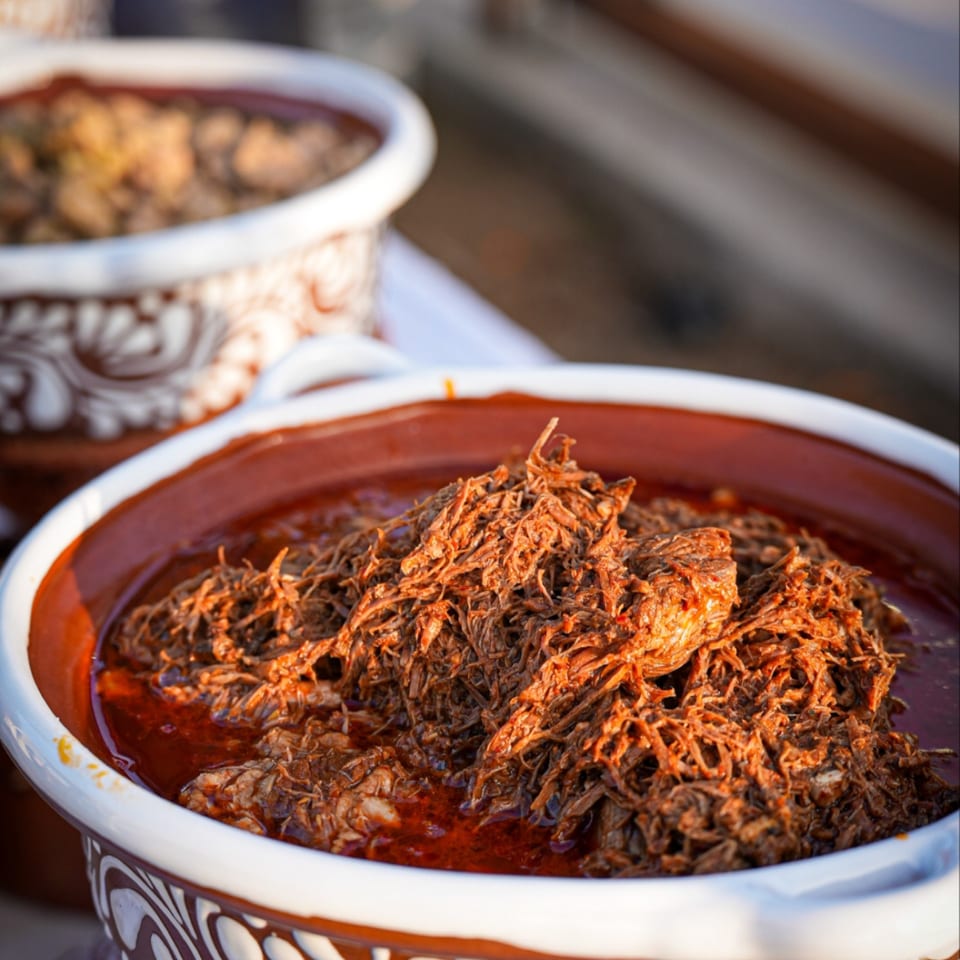 Close-up of a large ceramic bowl filled with shredded Mexican birria in a rich red sauce.