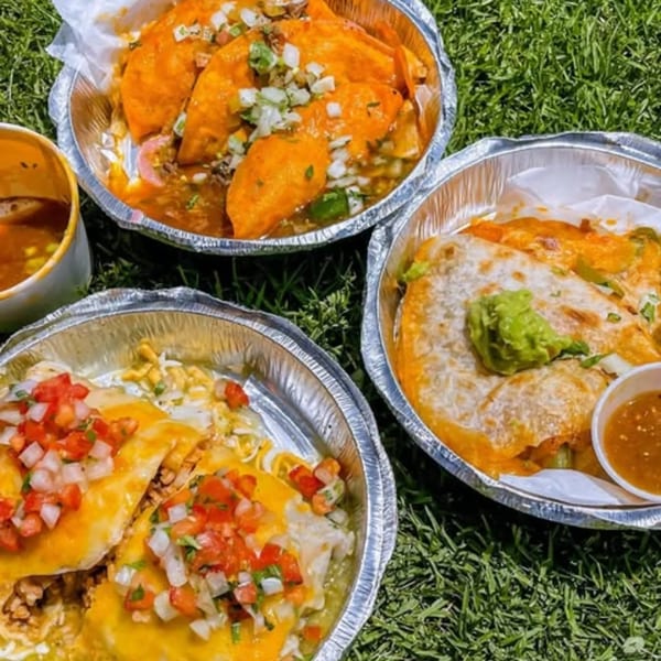Three foil containers of La Chancla Mexican Grill dishes, including tacos, quesadillas, and burritos, on grass.